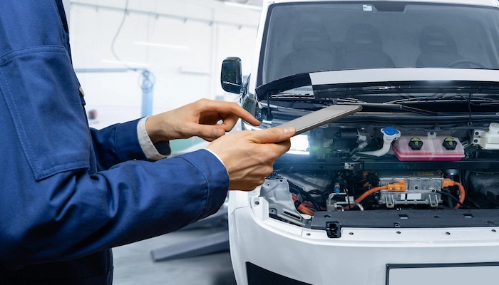 Technician using fleet maintenance software on a tablet while inspecting an electric vehicle engine compartment during a preventive maintenance check in a service workshop.